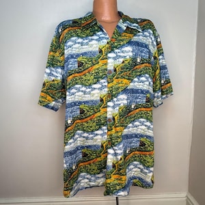 Puede incluir: Camisa de manga corta con botones y estampado de paisaje. La camisa presenta un patrón repetido de colinas verdes, cielo azul y nubes blancas. La camisa tiene un cuello clásico y botones de madera.