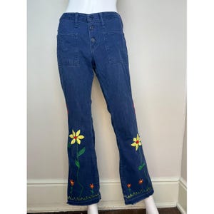 May include: Dark blue denim pants with embroidered floral design. The pants feature a button closure and two front pockets. Yellow, red, and orange flowers are embroidered along the lower legs, adding a colorful accent to the garment.