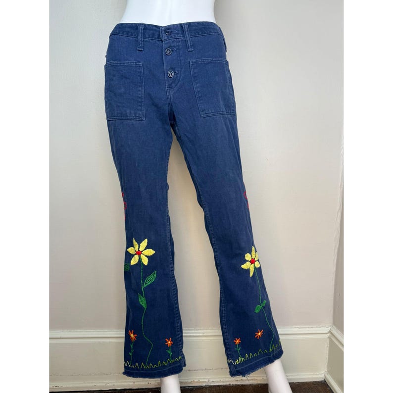 May include: Dark blue denim pants with embroidered floral design. The pants feature a button closure and two front pockets. Yellow, red, and orange flowers are embroidered along the lower legs, adding a colorful accent to the garment.