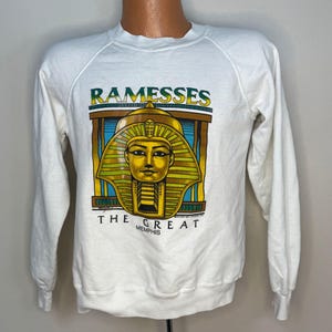 May include: White sweatshirt with a graphic of the pharaoh Tutankhamun. The graphic is in gold, green, blue, and brown, with the word "RAMESSES" above and "THE GREAT MEMPHIS" below.
