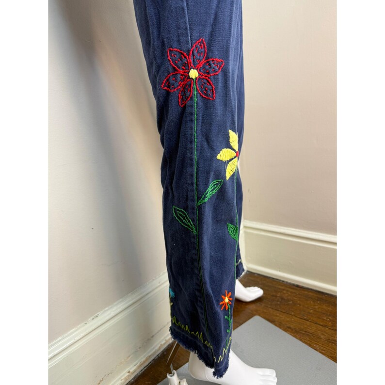 May include: Dark blue denim pants with colorful embroidered floral designs. The embroidery features red, yellow, and orange flowers with green stems and leaves. The pants have a frayed hem.