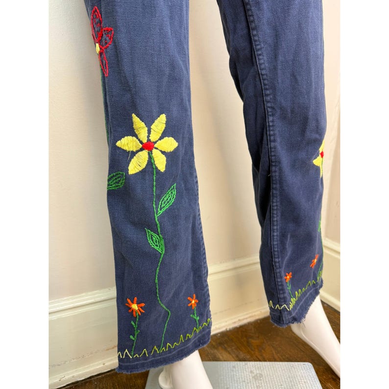 May include: Dark blue denim pants with embroidered floral designs. The embroidery features yellow, red, and orange flowers with green stems and leaves. The pants have a straight leg cut and a decorative hem.