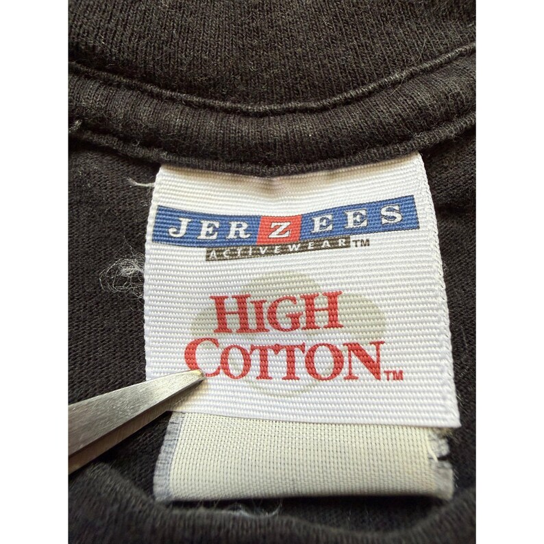 Pu&ograve; includere: Primo piano di un'etichetta di una maglietta nera. L'etichetta recita "JERZEES ACTIVEWEAR" in blu e bianco, con "HIGH COTTON" in rosso. Sono visibili un paio di forbici.
