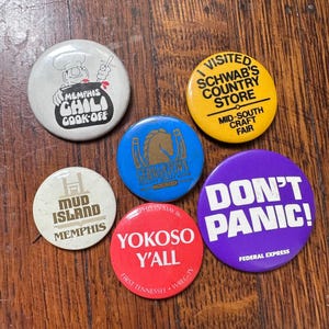 May include: Six round, colorful buttons with various designs and text. The buttons feature a chef with a pot of chili, a horse, a building with the text "Mud Island Memphis", a red button with the text "Yokoso Y'all", a purple button with the text "Don't Panic!" and a yellow button with the text "I Visited Schwab's Country Store Mid-South Craft Fair".