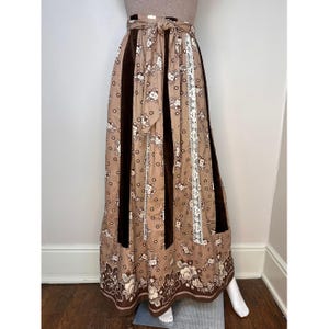 Pode incluir: Uma saia longa marrom e bege com estampa floral e uma faixa de veludo marrom na frente. A saia tem uma bainha de renda branca e um cinto marrom.