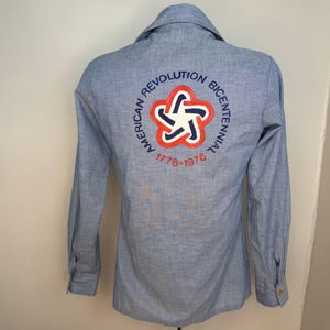 May include: Light blue button-down shirt with long sleeves. The back features an embroidered design with the text "AMERICAN REVOLUTION BICENTENNIAL 1776-1976" surrounding a red, white, and blue starburst emblem. The shirt has a collared neckline.