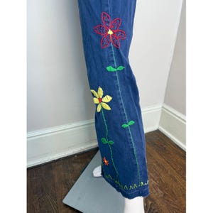May include: Blue denim pants with colorful embroidered flowers in red and yellow. Green stitching outlines the floral design and the pant's seams. The hemline has a decorative green zigzag stitch.