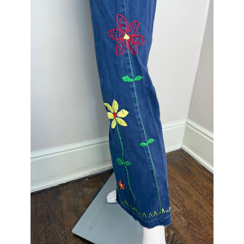 May include: Blue denim pants with colorful embroidered flowers in red and yellow. Green stitching outlines the floral design and the pant's seams. The hemline has a decorative green zigzag stitch.