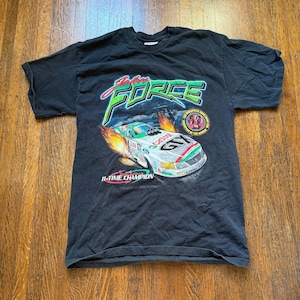 Pu&ograve; includere: T-shirt nero con grafica di un'auto da corsa e la scritta "John Force". Il design include la scritta "11-TIME CHAMPION" e un emblema circolare. L'auto &egrave; bianca, verde e rossa.