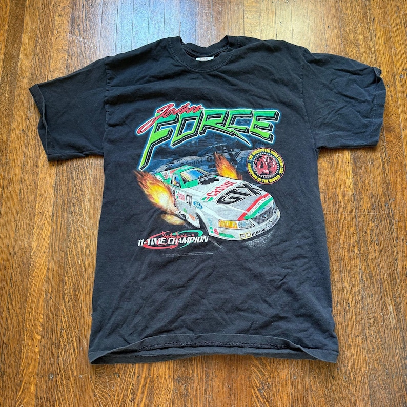 Pu&ograve; includere: T-shirt nero con grafica di un'auto da corsa e la scritta "John Force". Il design include la scritta "11-TIME CHAMPION" e un emblema circolare. L'auto &egrave; bianca, verde e rossa.