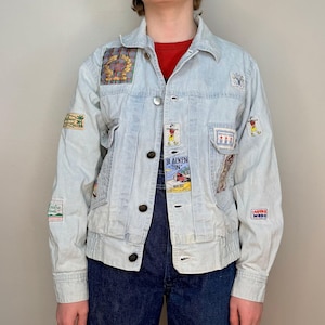 May include: Light blue denim jacket with various colourful patches, including a cartoon figure, a car, and a building. The jacket has a button closure and two pockets.