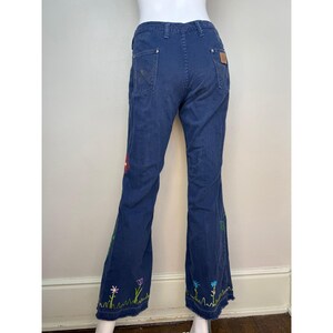 May include: Blue denim pants with a flared leg and embroidered floral design. The embroidery features colorful flowers and green grass along the bottom hem of the pants. The pants have a classic five-pocket design.