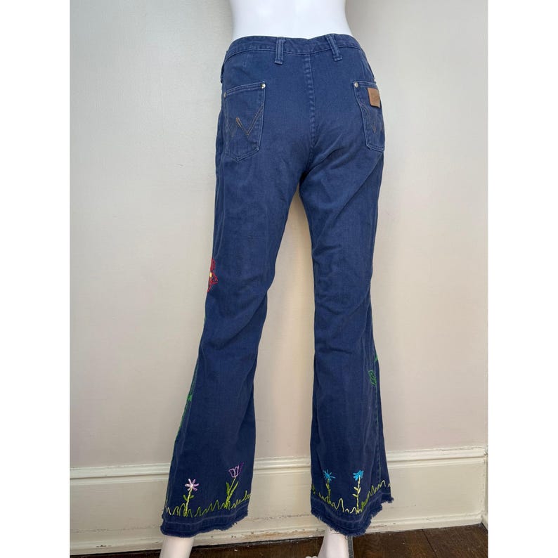 May include: Blue denim pants with a flared leg and embroidered floral design. The embroidery features colorful flowers and green grass along the bottom hem of the pants. The pants have a classic five-pocket design.