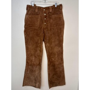 Könnte beinhalten: Braune Wildlederhose mit Knopfleiste und großen Taschen. Die Hose hat eine weite Beinweite.