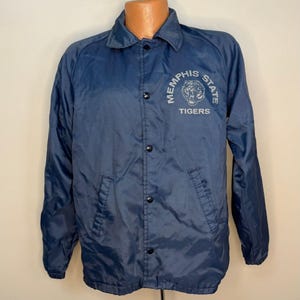 Puede incluir: Chaqueta de entrenador azul marino con cierre de botones y cuello. La chaqueta presenta el texto "MEMPHIS STATE TIGERS" en un diseño circular en el pecho. La chaqueta tiene dos bolsillos delanteros y mangas largas.