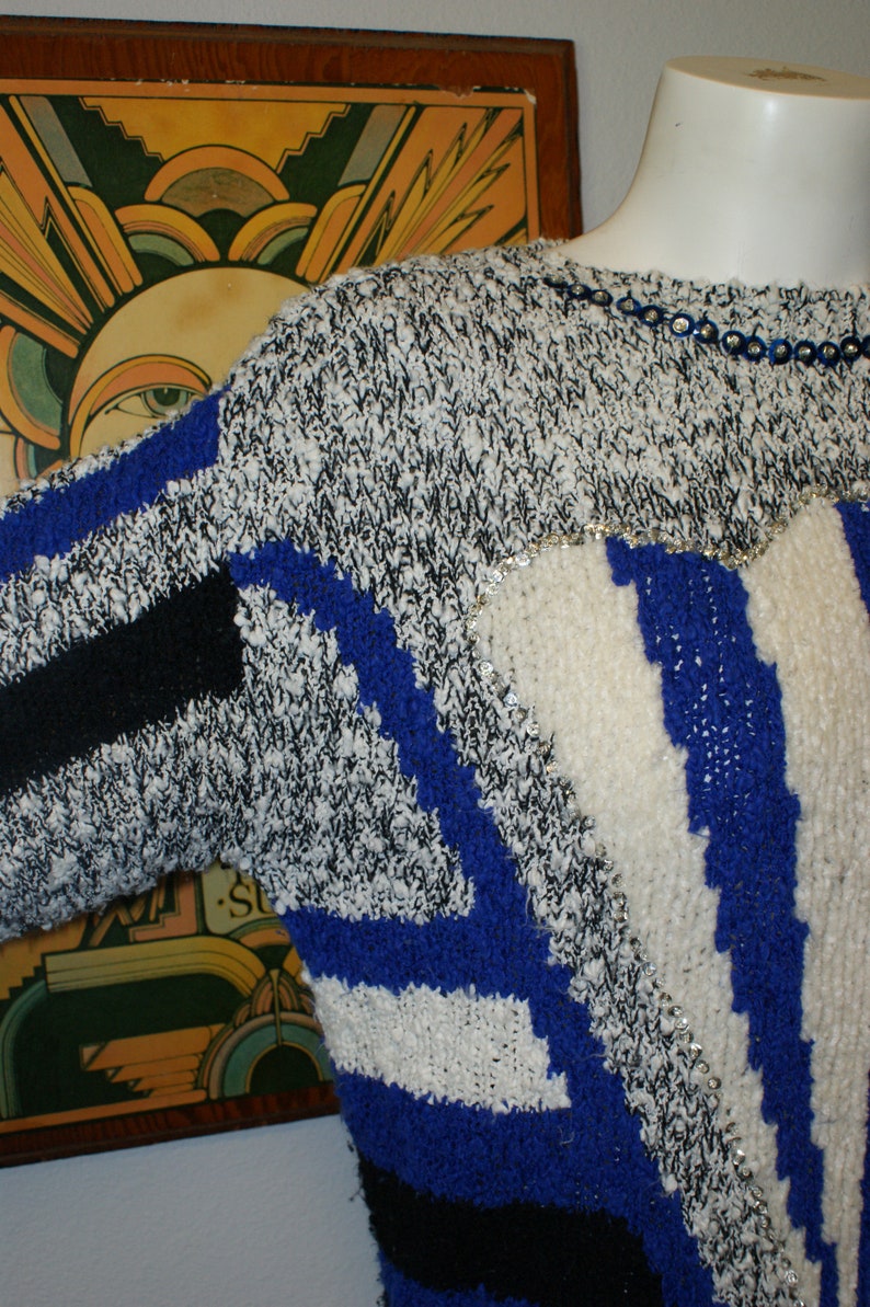 May include: A vintage sweater with a unique design. The sweater features a mix of black, white, and blue knit patterns, with a touch of silver sequins. The design includes stripes and geometric shapes, creating a bold and retro look.