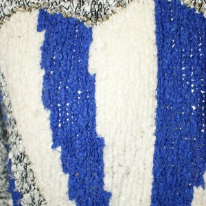 May include: Close-up of a textured sweater with a blue and white striped design. The top portion has a black and white knit pattern with silver accents. The blue stripes have a raised, textured appearance.