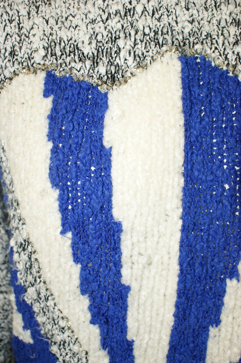 May include: Close-up of a textured sweater with a blue and white striped design. The top portion has a black and white knit pattern with silver accents. The blue stripes have a raised, textured appearance.