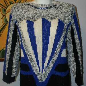 May include: A vintage sweater with a striking geometric design. The sweater features a mix of black, white, and blue knit patterns. The neckline is embellished with small, iridescent sequins. The overall style is reminiscent of the 1980s fashion.