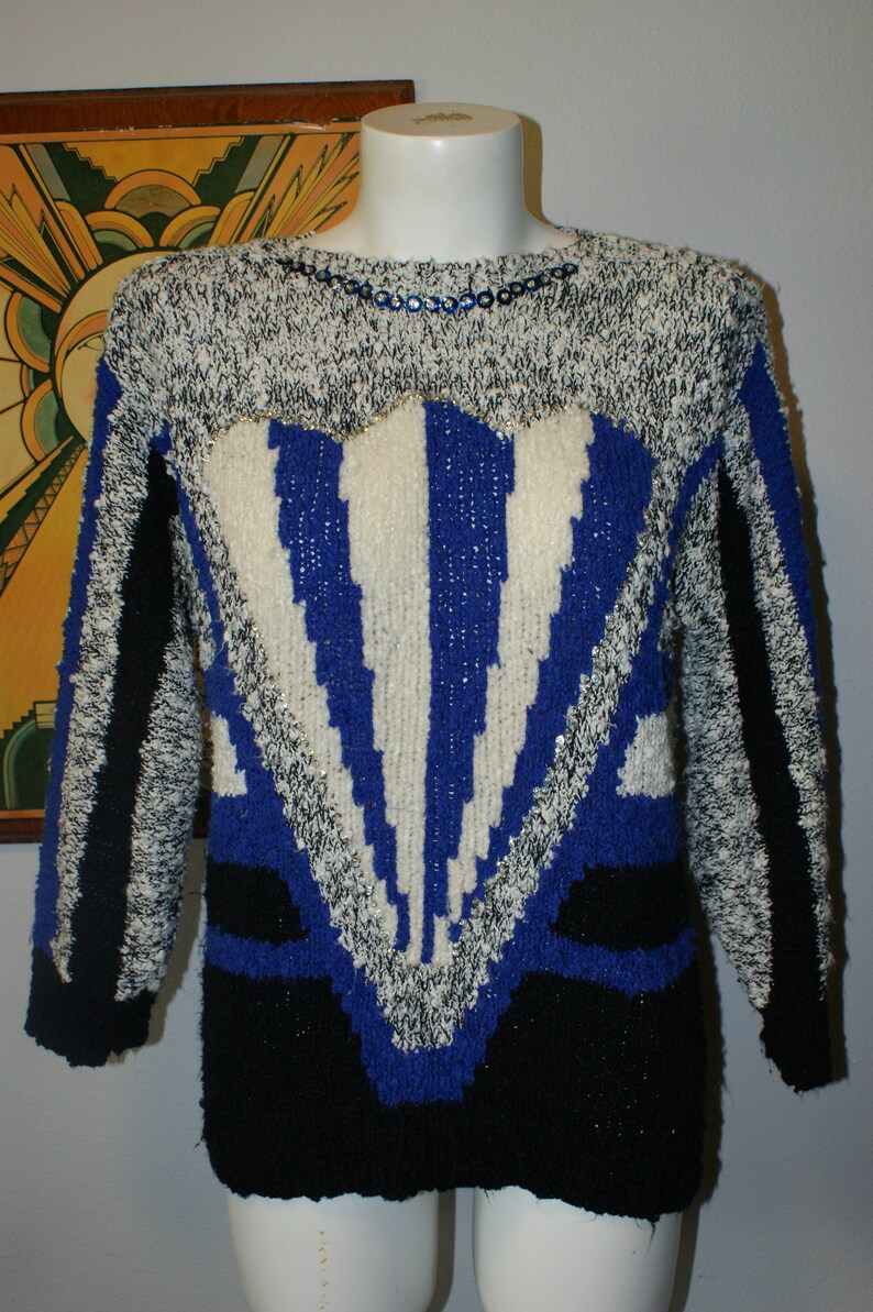 May include: A vintage sweater with a striking geometric design. The sweater features a mix of black, white, and blue knit patterns. The neckline is embellished with small, iridescent sequins. The overall style is reminiscent of the 1980s fashion.