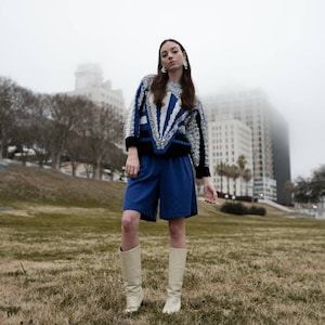 May include: A person wearing a blue and white patterned sweater, blue shorts, and cream-colored boots. The sweater has a geometric design with black accents. The outfit is set against a backdrop of a grassy area and buildings on a foggy day.