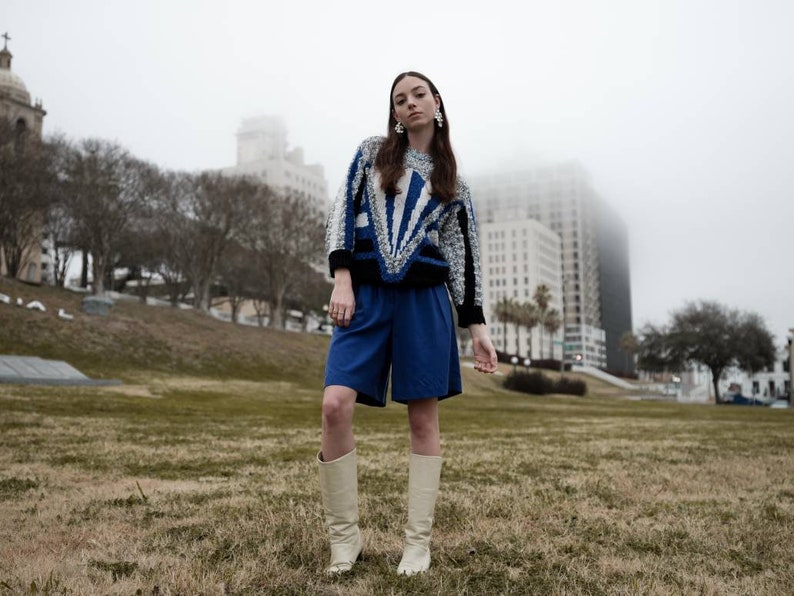 May include: A person wearing a blue and white patterned sweater, blue shorts, and cream-colored boots. The sweater has a geometric design with black accents. The outfit is set against a backdrop of a grassy area and buildings on a foggy day.