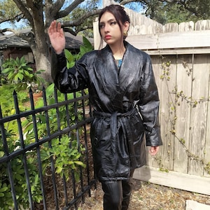 May include: Black leather coat with a tie belt, worn with matching leather pants. The coat has a quilted design and a wide collar. The person is standing outside near a black metal fence and a wooden fence.