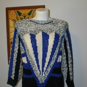 May include: A vintage sweater with a striking geometric design. The sweater features a mix of black, white, and blue colors. The upper portion is speckled, with a blue and white pattern in the center. The neckline is adorned with blue sequins.