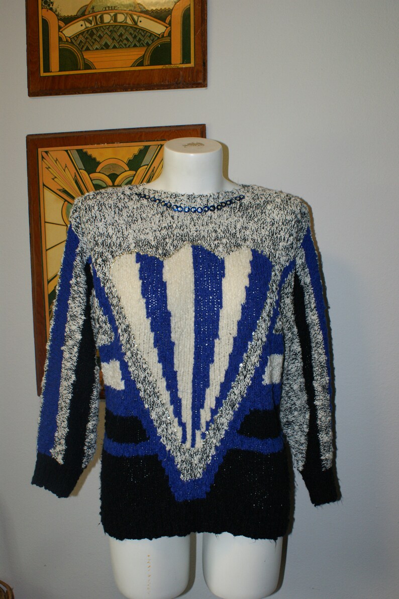 May include: A vintage sweater with a striking geometric design. The sweater features a mix of black, white, and blue colors. The upper portion is speckled, with a blue and white pattern in the center. The neckline is adorned with blue sequins.