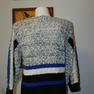 May include: A vintage sweater with a speckled gray and white knit pattern. The sweater features blue and black stripes across the midsection and on the sleeves. The cuffs and lower hem are black.