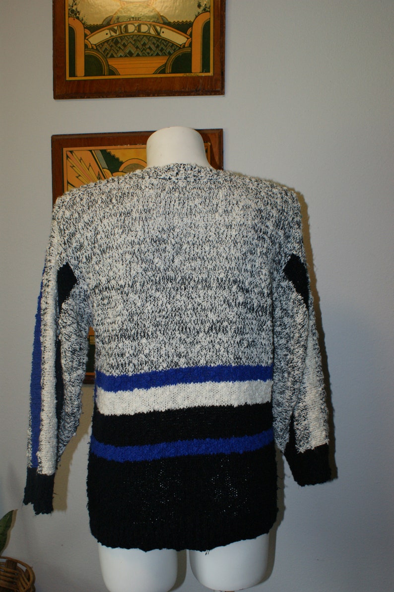 May include: A vintage sweater with a speckled gray and white knit pattern. The sweater features blue and black stripes across the midsection and on the sleeves. The cuffs and lower hem are black.