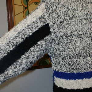 May include: A textured sweater with a black and white speckled pattern. The sleeve has a black stripe. The bottom of the sweater has horizontal stripes of blue, white, and black. The sweater is made of a thick, possibly wool, material.