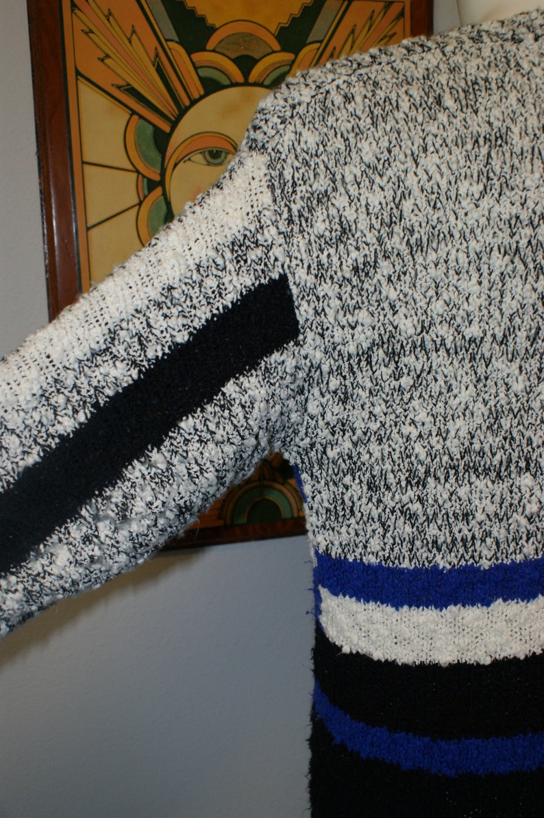 May include: A textured sweater with a black and white speckled pattern. The sleeve has a black stripe. The bottom of the sweater has horizontal stripes of blue, white, and black. The sweater is made of a thick, possibly wool, material.