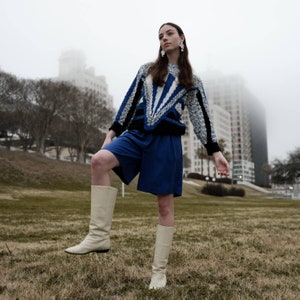 May include: A person wearing a blue and white patterned sweater, blue shorts, and cream-colored boots stands on a grassy area. The sweater has a geometric design. The background includes a city skyline and a foggy sky.
