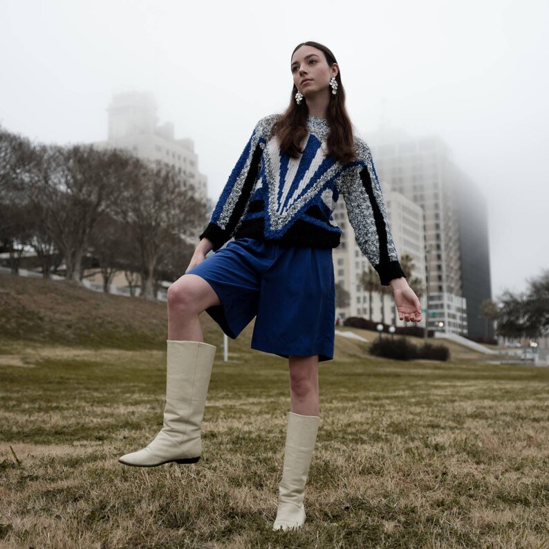 May include: A person wearing a blue and white patterned sweater, blue shorts, and cream-colored boots stands on a grassy area. The sweater has a geometric design. The background includes a city skyline and a foggy sky.