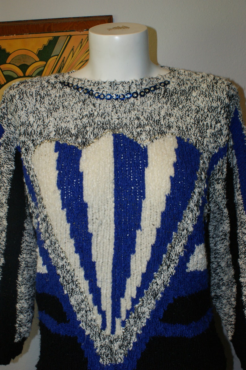 May include: A vintage sweater with a unique design. The sweater features a heart-shaped pattern with blue and white stripes, outlined with silver thread. The upper portion is a mix of black and white knit, with blue sequin detailing around the neckline.