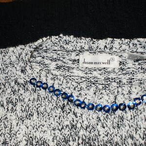 May include: A close-up of a white and black knit sweater with a crew neck. The sweater has a row of blue sequins along the neckline. The label reads "dason maxwell".