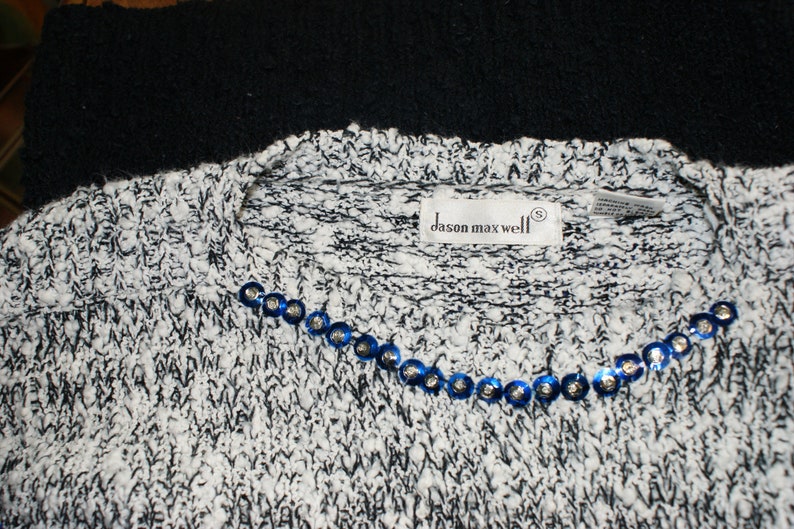 May include: A close-up of a white and black knit sweater with a crew neck. The sweater has a row of blue sequins along the neckline. The label reads "dason maxwell".