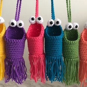 May include: Six crocheted hanging baskets with googly eyes and fringe. The baskets are yellow, purple, pink, blue, green, and orange.