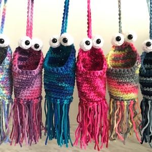 May include: Six crocheted hanging baskets with colourful yarn and googly eyes. The baskets have fringe at the bottom and are made in a variety of colours, including pink, blue, purple, and green.