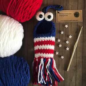 May include: A handmade crochet hanging basket in red, white, and blue, resembling a character with large googly eyes. The basket has a striped pattern and fringe. A crochet hook, yarn, and star-shaped embellishments are also present.
