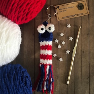 May include: A patriotic crochet keychain in red, white, and blue, resembling a rocket with googly eyes. The keychain is next to a crochet hook, star-shaped embellishments, and balls of yarn. The tag reads "Mini Yip Yip Martians Key Chain".