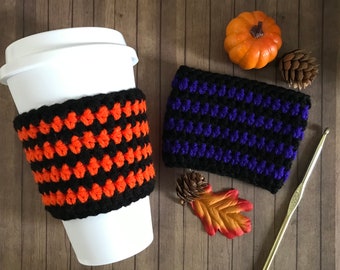 May include: A white reusable coffee cup featuring a black and orange crocheted sleeve, alongside a separate black and purple crocheted sleeve, a small orange pumpkin, two pinecones, an autumn leaf, and a gold-colored metal crochet hook.