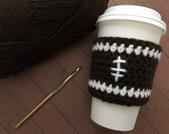 May include: A brown crocheted coffee cup cozy with white accents, designed to resemble a football. A ball of brown yarn and a gold crochet hook are also in the image. The cozy is wrapped around a white paper cup.