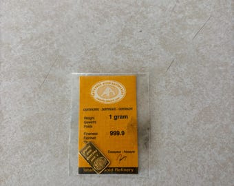 May include: A small gold bar, weighing 1 gram, is displayed on a yellow certificate. The certificate includes text in multiple languages, indicating the bar's fineness of 999.9. The gold bar is from Istanbul Gold Refinery.