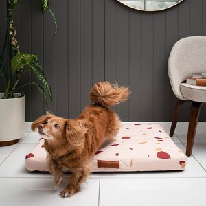 May include: A brown dog with a fluffy tail is lying on a pink and brown patterned dog bed. The bed has a brown leather tag with the word "Bark" on it.