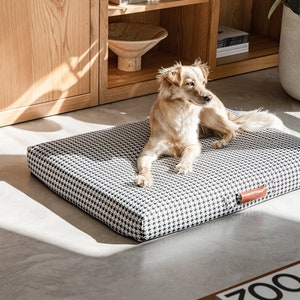 May include: A light brown dog laying on a black and white checkered dog bed with a brown leather tag. The bed is on a light gray floor.