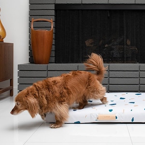 May include: A brown and white dog bed with a terrazzo pattern. The bed is on a white tiled floor in a living room with a fireplace and a black leather armchair.
