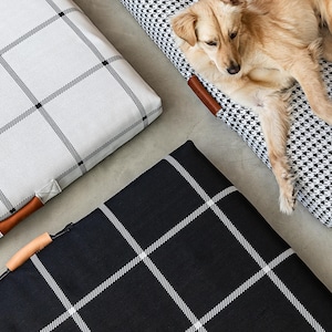 May include: Dog beds in black and white houndstooth and plaid patterns. One bed is occupied by a light brown dog. The beds have brown leather handles and are on a gray surface. The plaid beds are white and black.