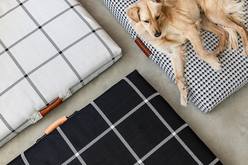 May include: Dog beds in black and white houndstooth and plaid patterns. One bed is occupied by a light brown dog. The beds have brown leather handles and are on a gray surface. The plaid beds are white and black.